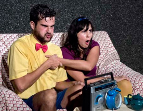 imagen de la representación: dos personajes en un sofá miran la TV sorprendidos